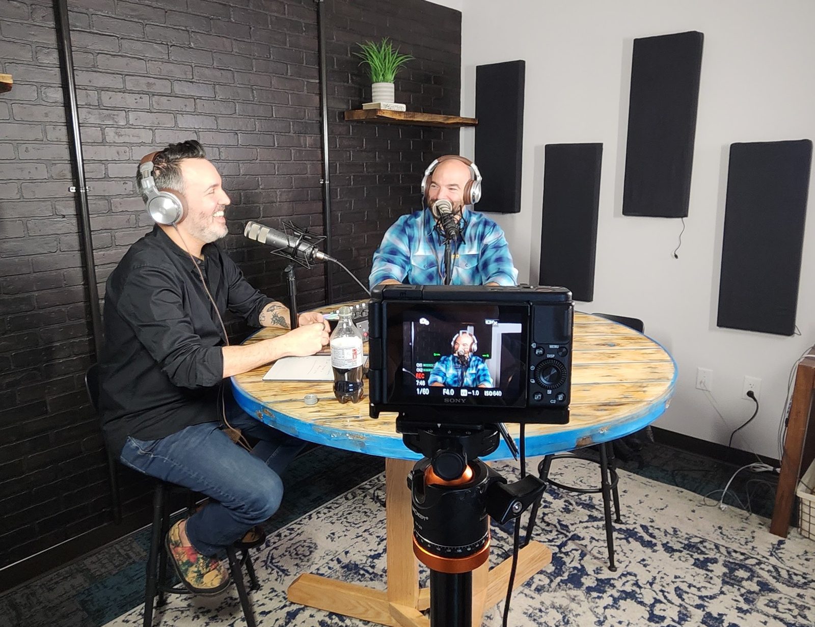 Two podcast hosts recording at a studio table with a camera in the foreground